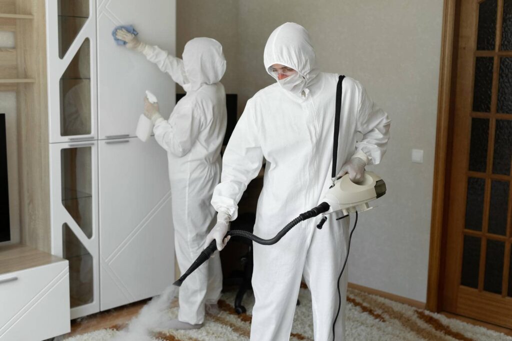 Two people in white protective suits disinfect a room, one spraying steam and the other wiping a cabinet.