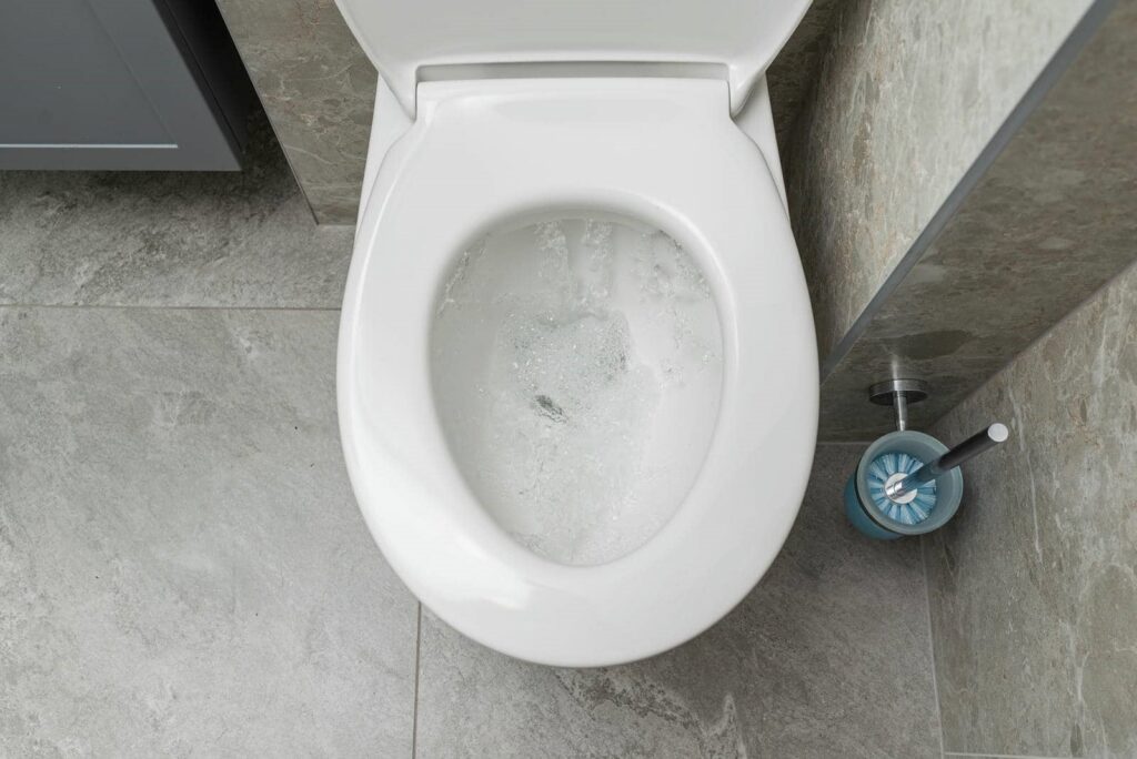 White toilet flushing in a bathroom with gray tiled floor and a blue toilet brush holder.